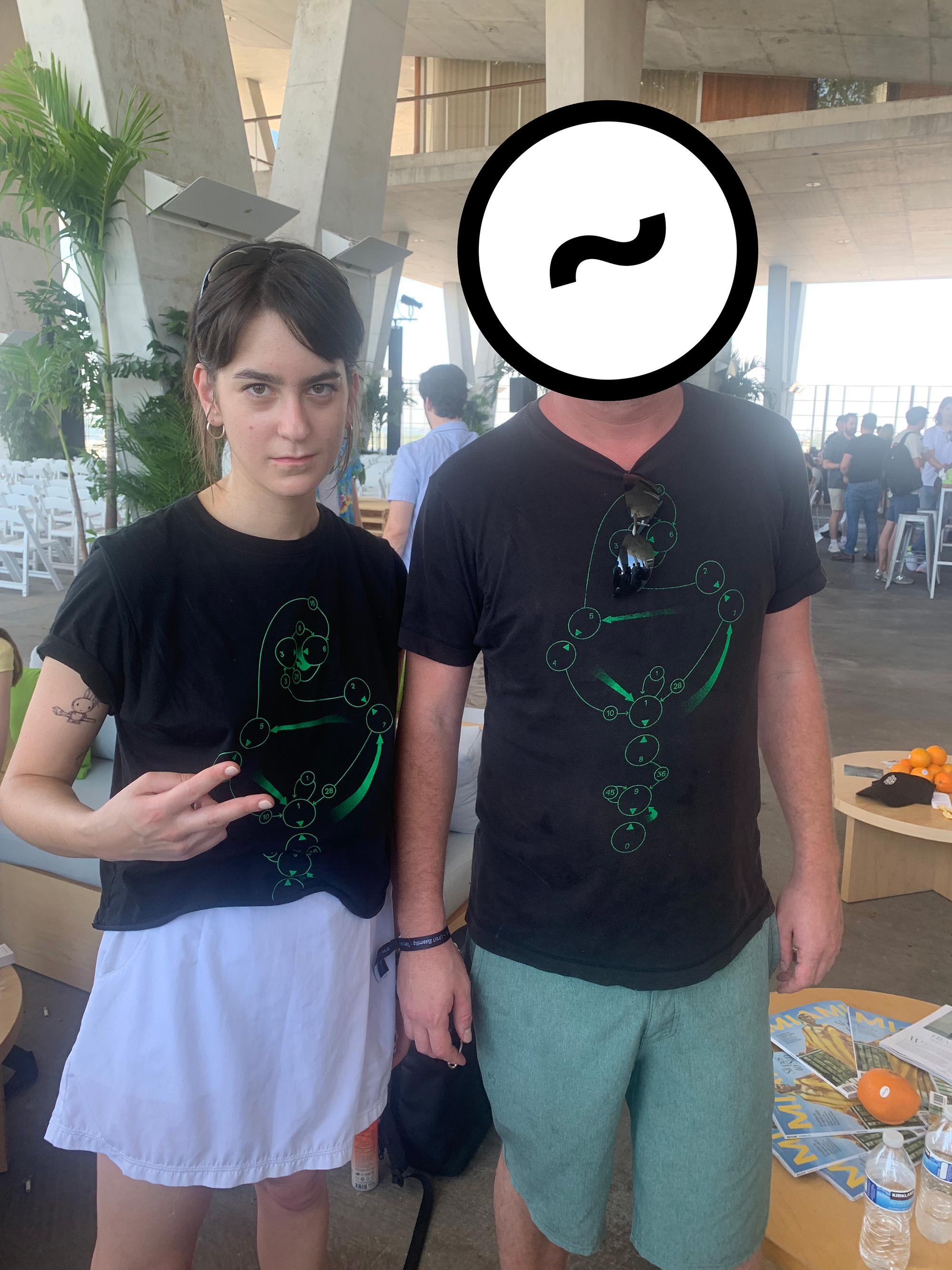 two people - a woman with dark hair and a man whose face is obscured by an urbit logo - wear matching t-shirts bearing the numogram symbol designed by the cybernetic culture research unit