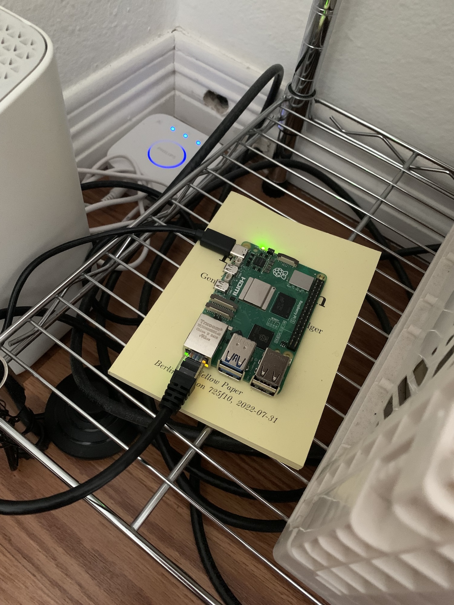a green raspberry pi sitting atop a small yellow book on a silver wire shelf, with black cords protruding in several directions.