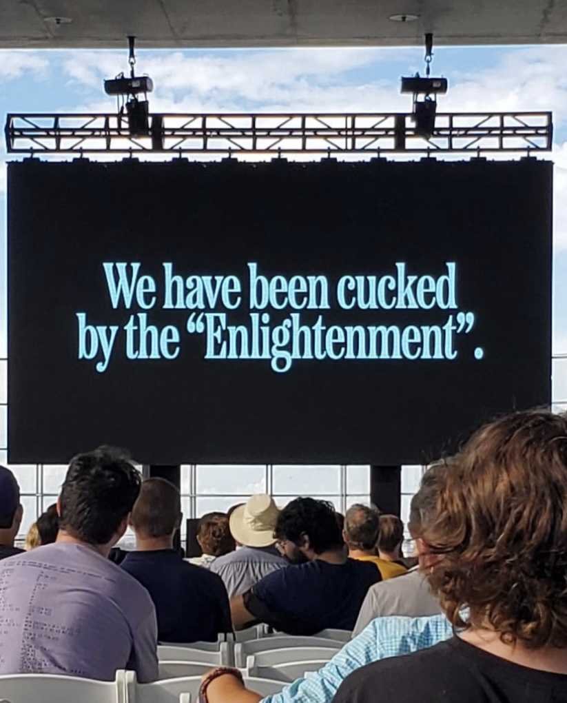people sitting in folding chairs facing a stage are visible from behind. they look at a screen. white text on the black screen reads 'we have been cucked by the enlightenment'
