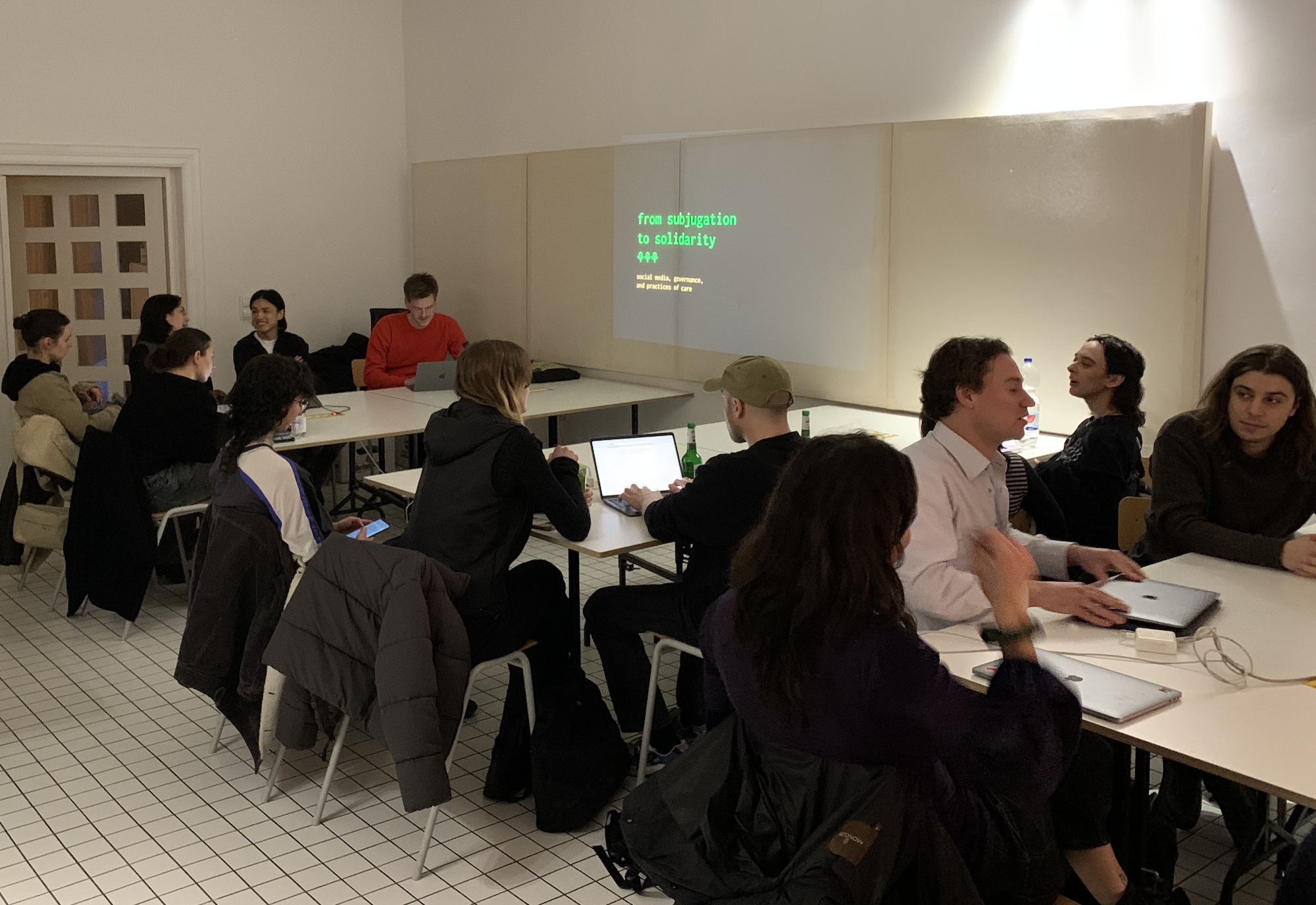 groups of people at laptops are clustered around tables near a projected slide that reads 'from subjugation to solidarity.' they are in a room with white walls and a white tile floor.