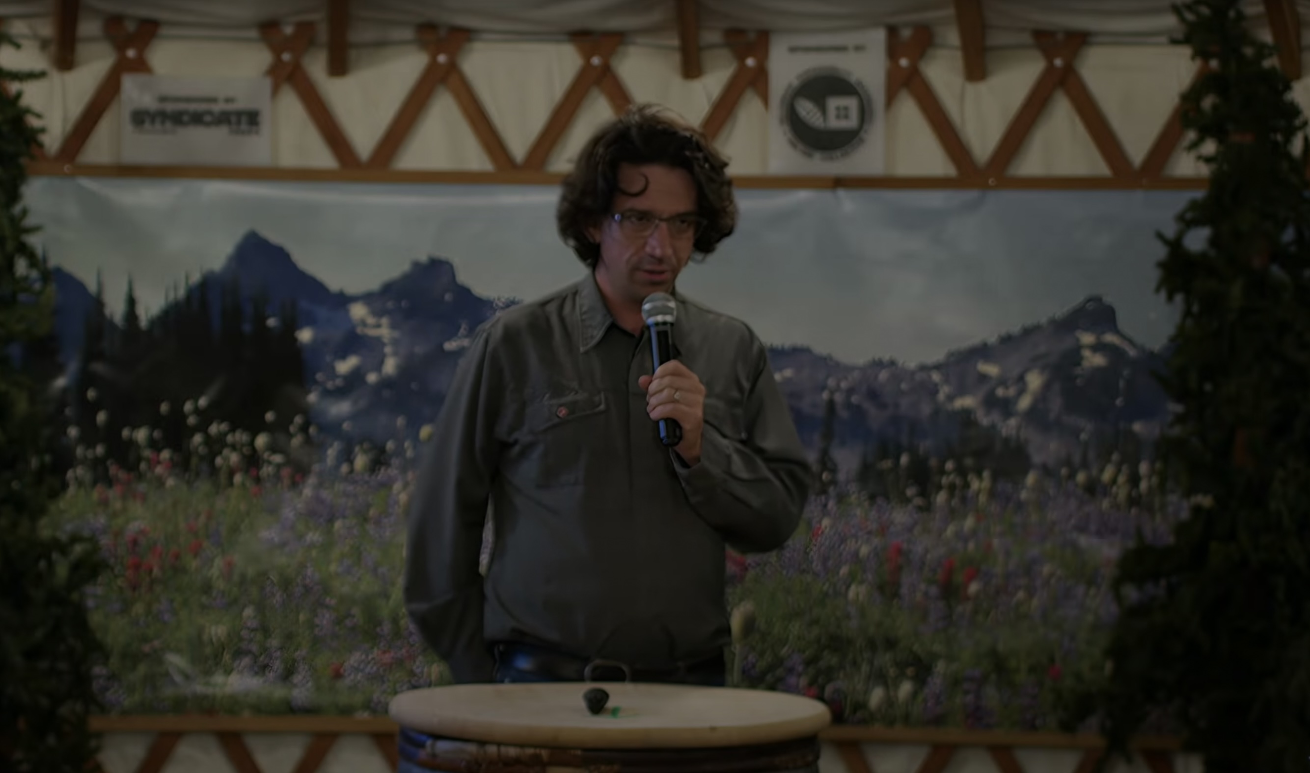 a middle-aged white man with floppy brown hair speaks into a handheld microphone. a single lock of curly hair is cartoonishly plastered to his sweaty face. he wears an olive-green button down and stands in front of a mural depicting a valley of wildflowers and a rolling mountain range.